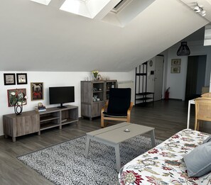 A modern living room with a flat-screen TV, wooden furniture, a patterned rug, and a loft-style ceiling.