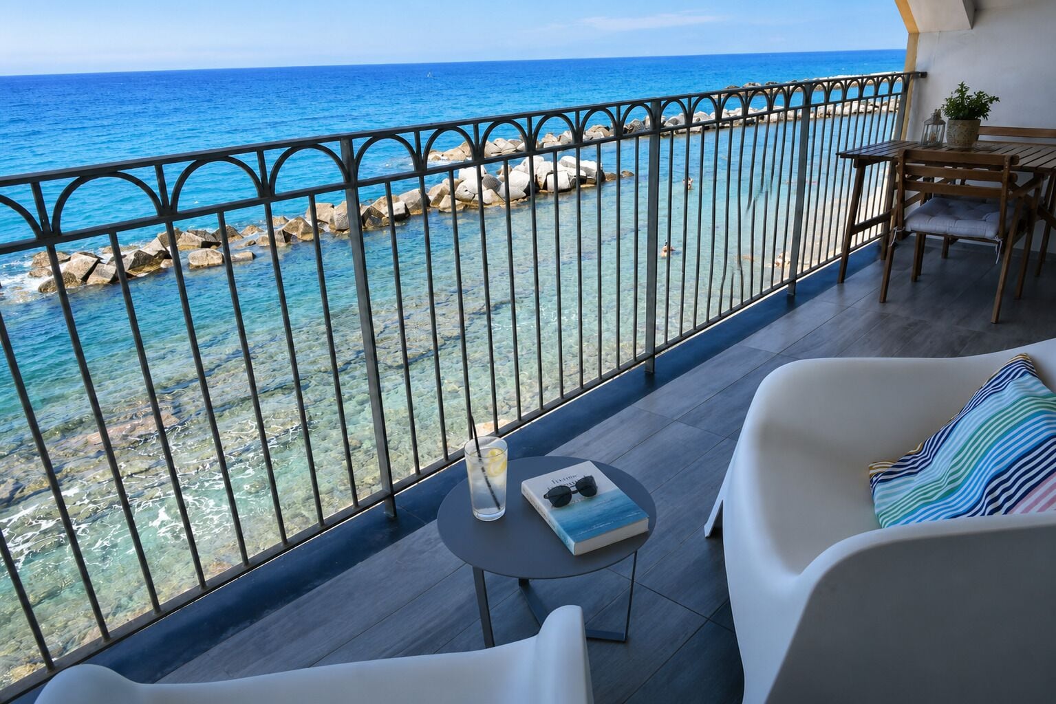 Un balcon avec vue sur la mer, doté d’une balustrade en métal, d’une table avec un livre et des lunettes, et de deux chaises blanches.