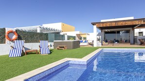 A swimming pool with a tiled edge, surrounded by a grassy area with two striped lounge chairs and a hot tub.