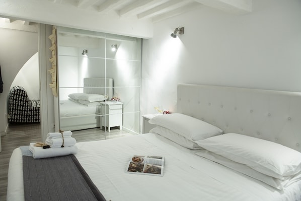 A modern hotel room with a large bed, a glass partition, and a small bedside table with a lamp.