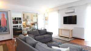 A living room with a grey sofa set, a television mounted on the wall, and a kitchen area visible through an open door. - Family House (Cefalù)
