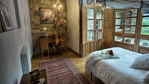 A bedroom with a large bed, a desk with a chair, a stone wall, and a window with a view of greenery.
