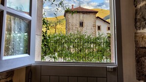 A window view of a rustic building with a tiled roof, surrounded by greenery and a mountainous backdrop.