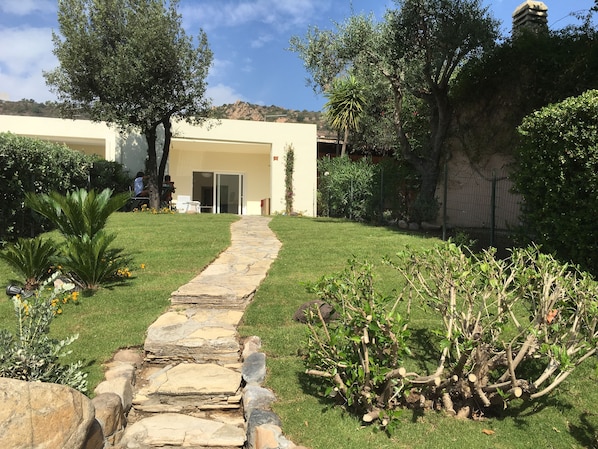 A stone pathway leading to a building with a glass door, surrounded by greenery and a stone wall.