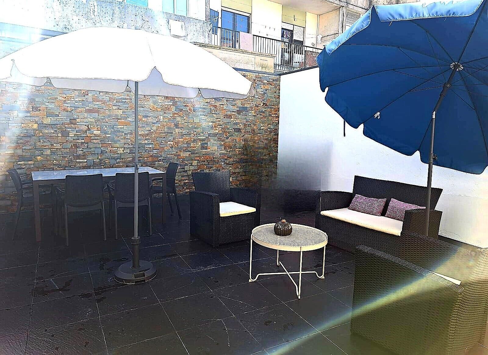 Un patio al aire libre con una pared de piedra, una mesa de comedor con sillas, una mesa redonda con una lámpara y dos sofás con sombrillas.