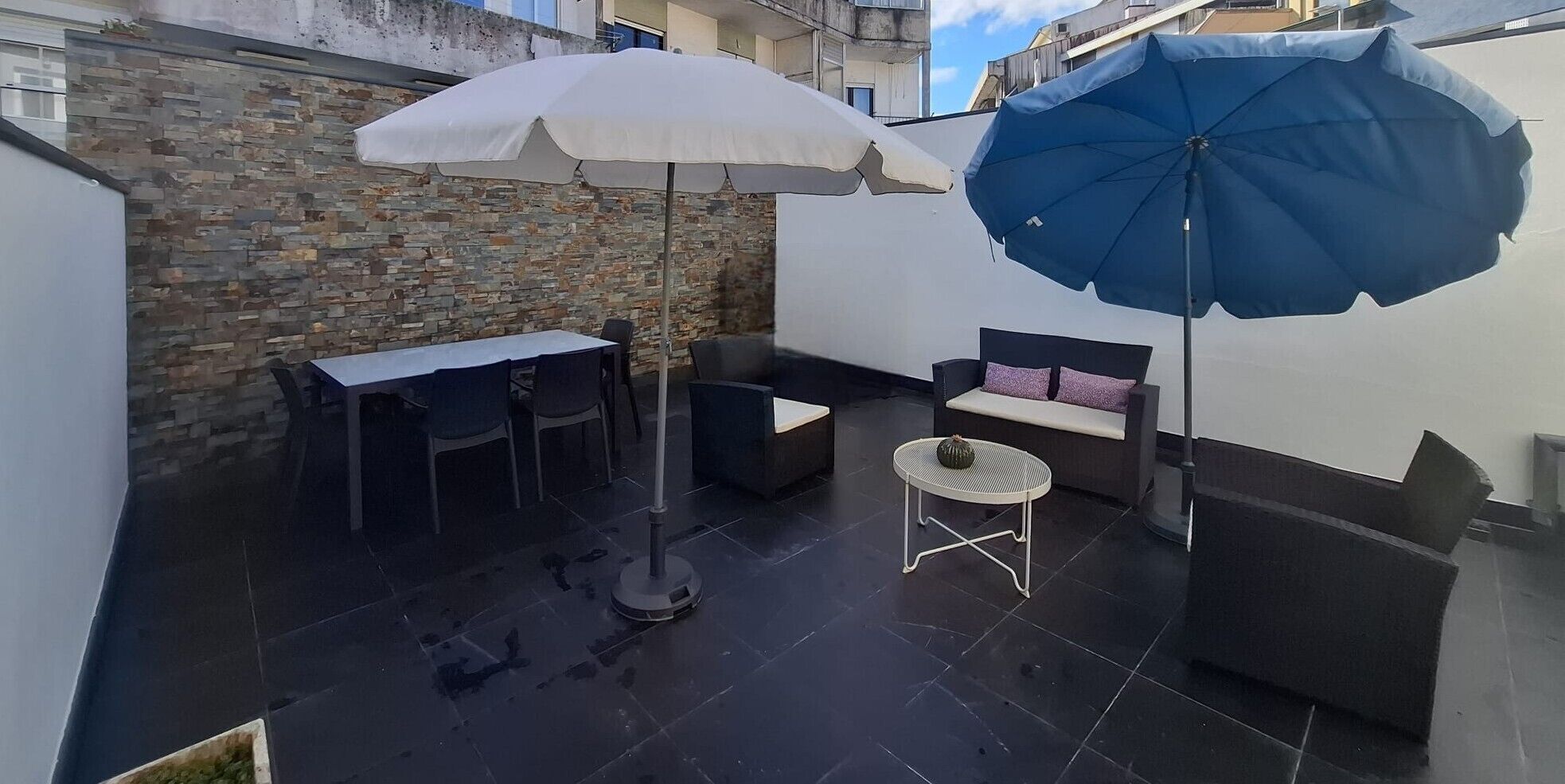 En tagterrasse med stenvæg, et bord med stole, en sofa og to parasoller.