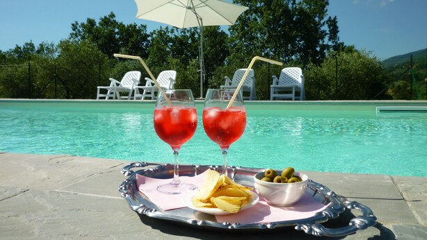 Cenário à beira da piscina com duas bebidas, uma bandeja de petiscos e um guarda-sol branco.