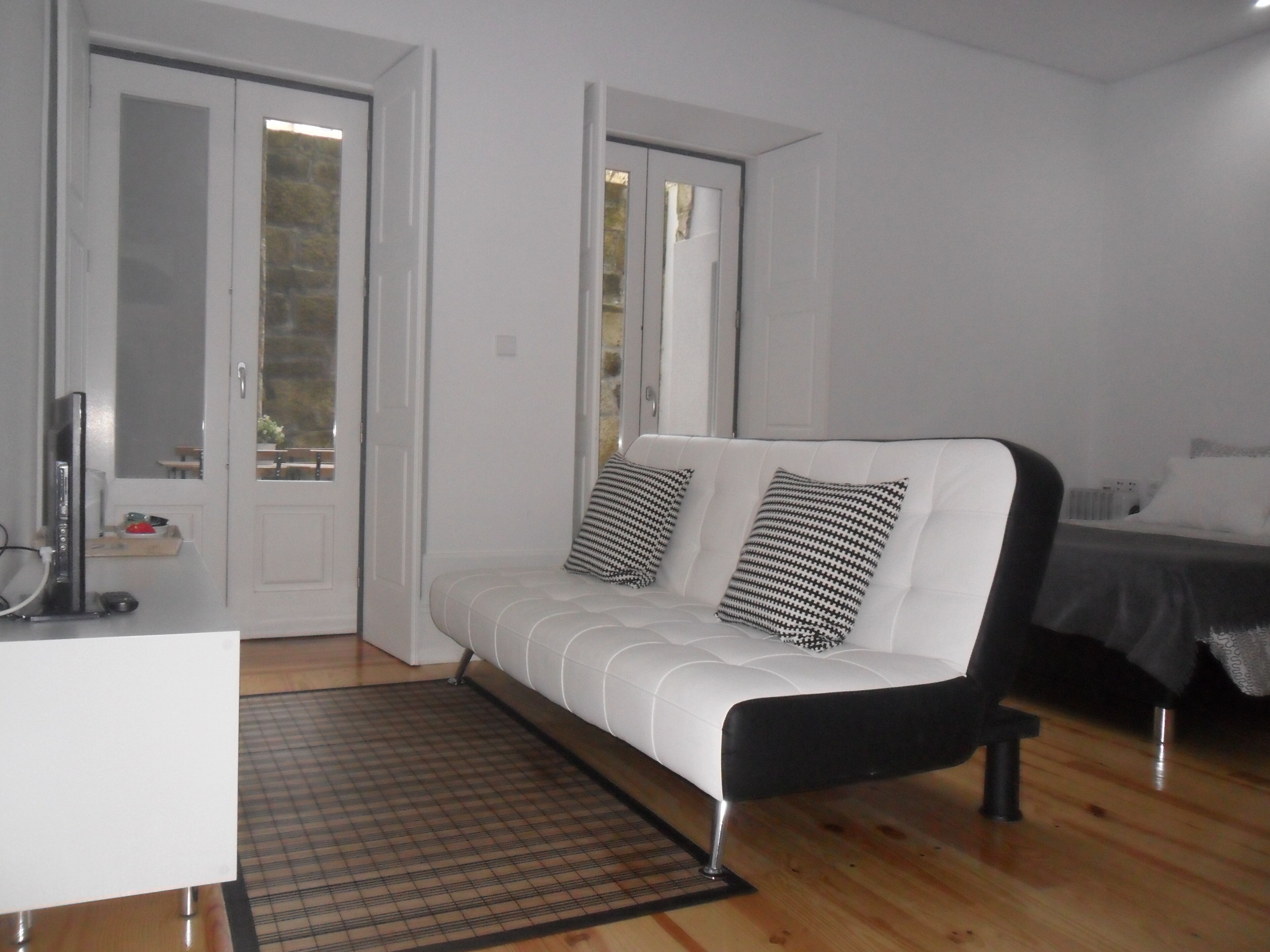 A modern, minimalist bedroom with a white sofa, two black and white patterned pillows, a wooden floor, and a small desk with a computer.