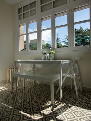 Una habitación con una mesa y sillas blancas, suelo estampado y ventana con vista a árboles y edificios.