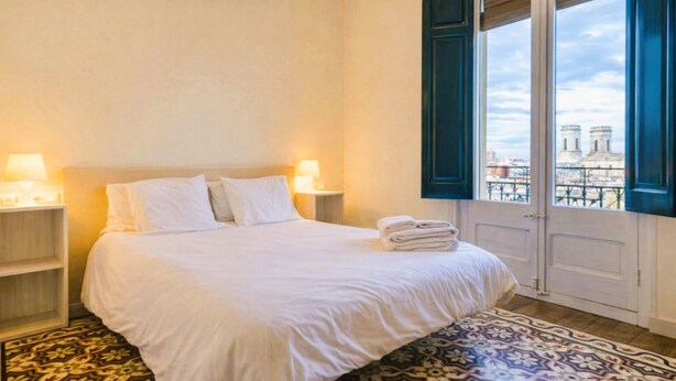 A hotel room with a bed, nightstands, a patterned rug, and a view of a building through a window.