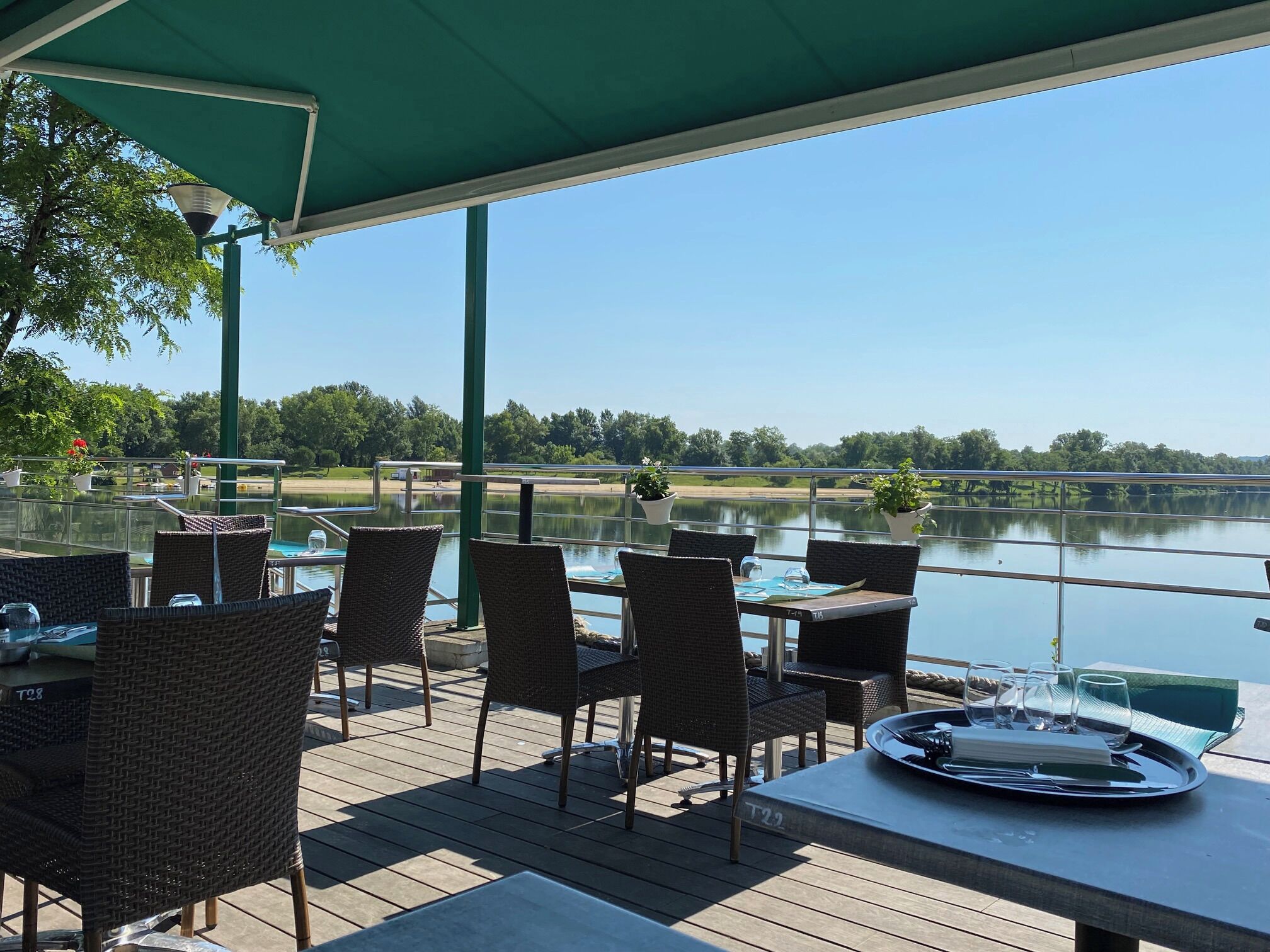 Un espacio comedor al aire libre con techado, sillas y mesas de mimbre, con vista a un lago tranquilo y un puente.
