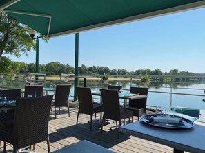 Een overdekt terras met rieten stoelen en tafels, met uitzicht op een rustig meer en een brug.