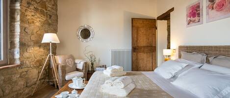 Una habitación acogedora con pared de piedra, techo de madera, cama con sábanas blancas, una silla de mimbre, una mesa con juego de té y un espejo en la pared.
