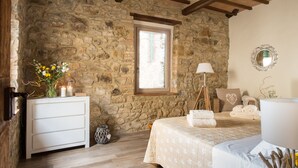 A bedroom with stone walls, a wooden ceiling, a bed with a quilt, a bedside table with a lamp, and a window.