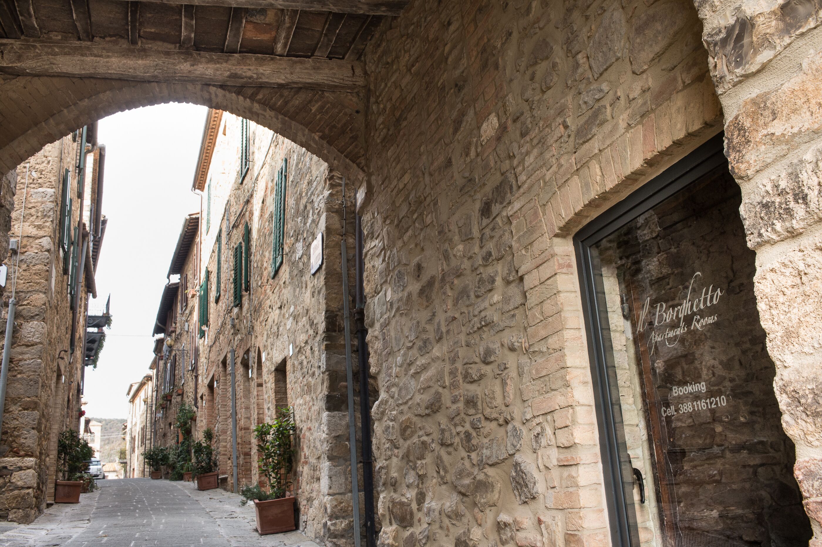 Une rue étroite bordée d’immeubles en pierre, un arc de triomphe et une porte sur laquelle est apposée une enseigne indiquant « M. Borghetto ».