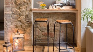 A rustic kitchen with stone walls, wooden beams, a bar area with stools, and a small table with kitchenware and flowers.