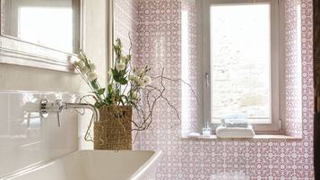 A bathroom with a large bathtub, a small stool with towels, a wicker basket, and a window with a view.