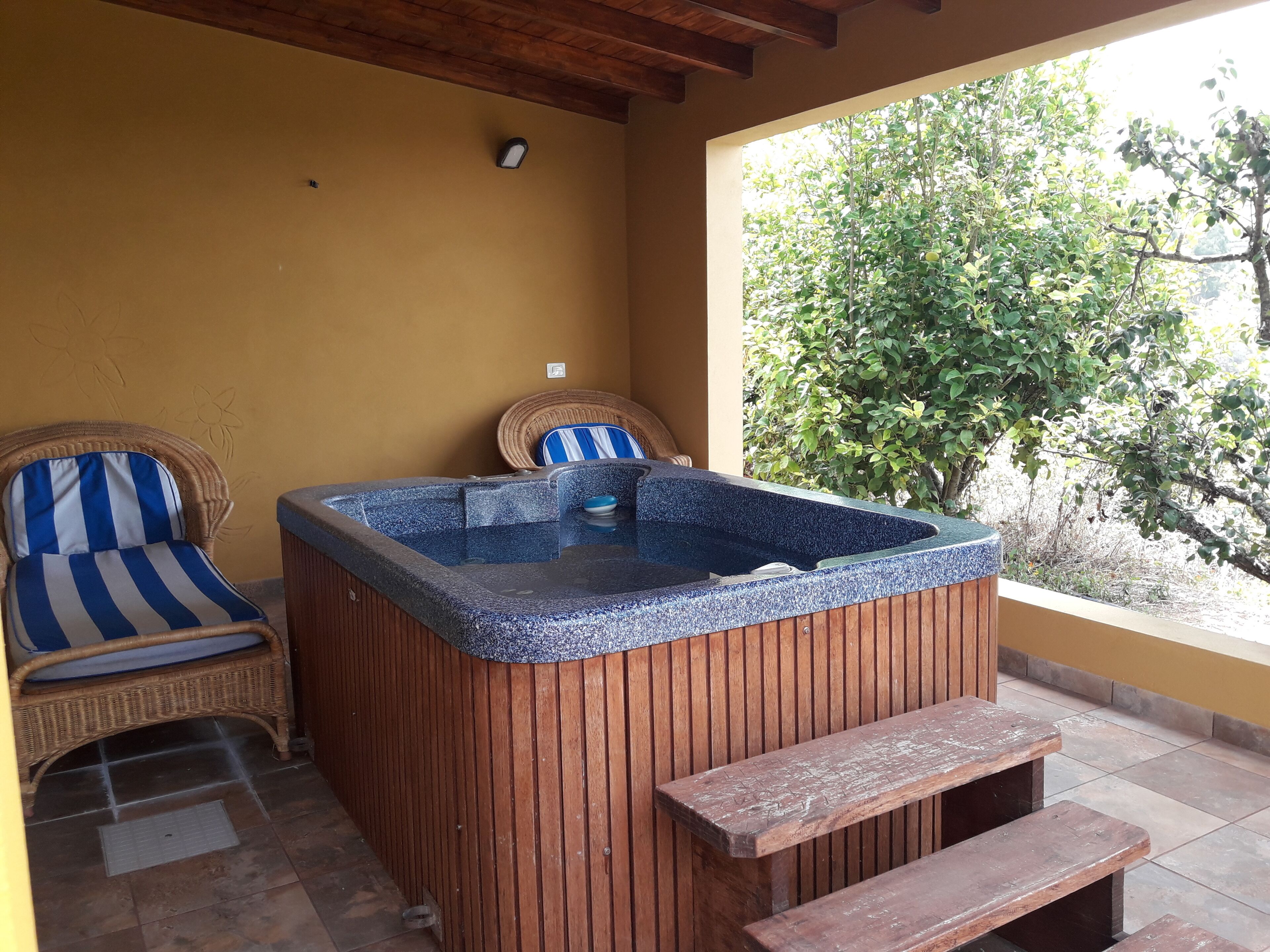A hot tub with wooden benches, a wicker chair, and a view of greenery.