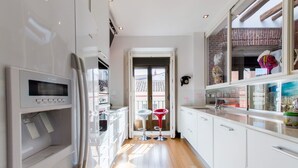 Une cuisine moderne avec des armoires blanches, un four encastré et un réfrigérateur. On y trouve deux tabourets et une fenêtre donnant sur un bâtiment à l’extérieur.