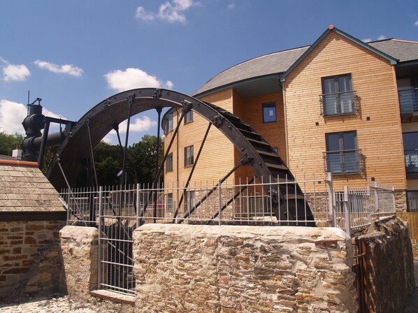A historic building with a large water wheel, stone walls, and a metal fence.