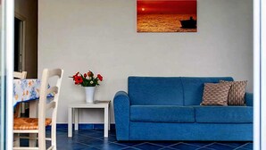 Een woonkamer met een blauwe bank, een kleine tafel met een bloevas en een aan de muur bevestigd schilderij van een zonsondergang boven de oceaan.