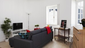 Ett modernt vardagsrum med en grå soffa, en platt-TV, ett litet bord med en växt och ett fönster med persienner.