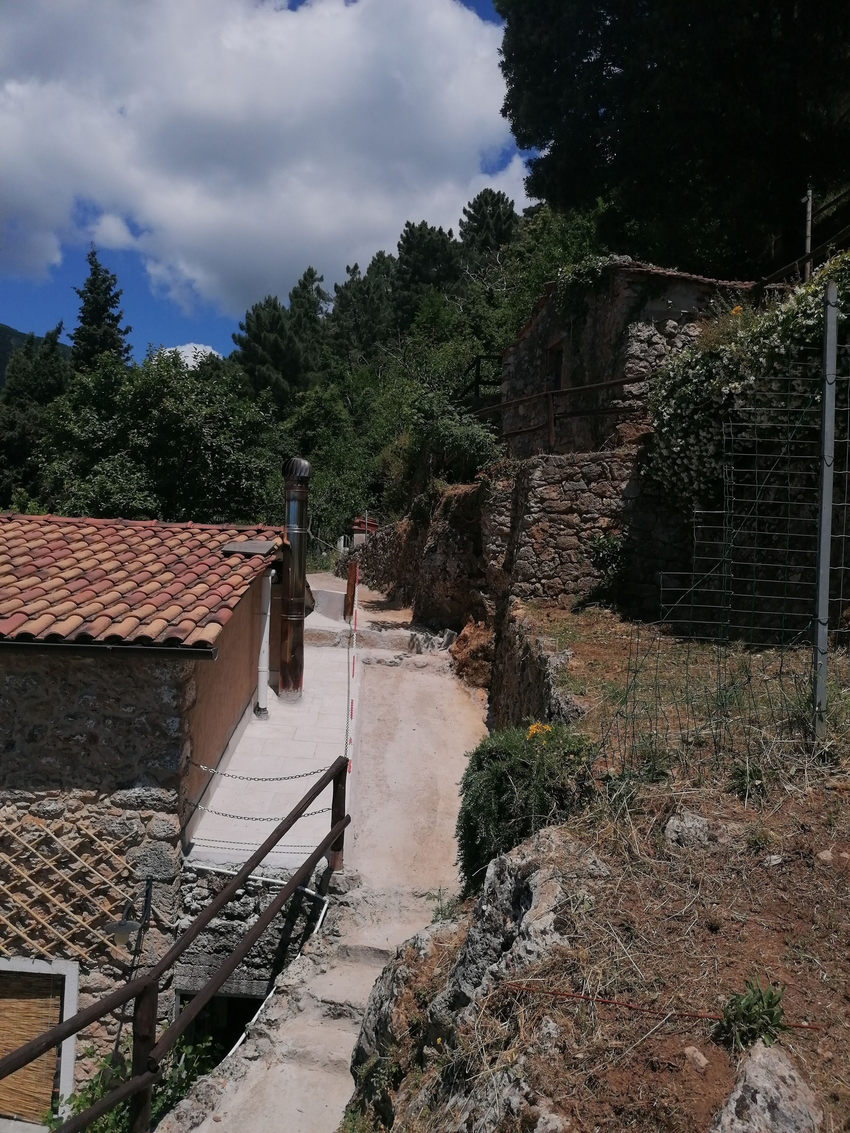 A narrow stone pathway leading to a traditional building with a tiled roof, surrounded by greenery and a stone wall.