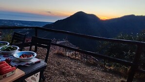 A table set outdoors with food and drinks, overlooking a mountainous landscape at sunset.