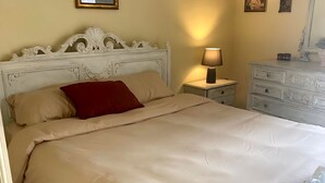 A bed with a white headboard, a red pillow, and a beige bedspread. There is a nightstand with a lamp and a dresser with a mirror.