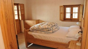Un lit en bois recouvert d’une couverture à motifs, une table de chevet avec une lampe et une fenêtre avec des volets en bois.