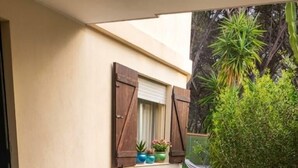 Un espace de restauration en plein air avec une table dressée pour quatre personnes, entourée de chaises blanches, d’une fenêtre avec des volets et de plantes en pot.