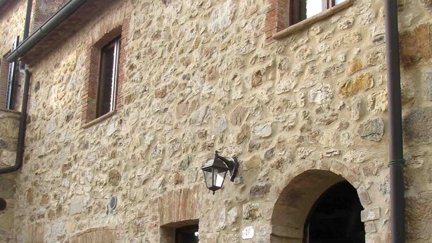 Edifici de pedra amb diverses finestres i una porta, que destaca un petit porxo amb plantes en testos.