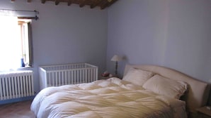Un dormitori amb un llit gran, un radiador blanc, una nitzeta amb una làmpada i una finestra amb cortina.