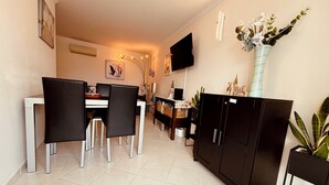 Een moderne eetruimte met een witte tafel, zwarte stoelen, een zwarte kast en een aan de muur bevestigde televisie.