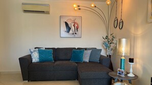 Een moderne woonkamer met een hoekbank, een klein rond tafeltje en een wandairconditioner.