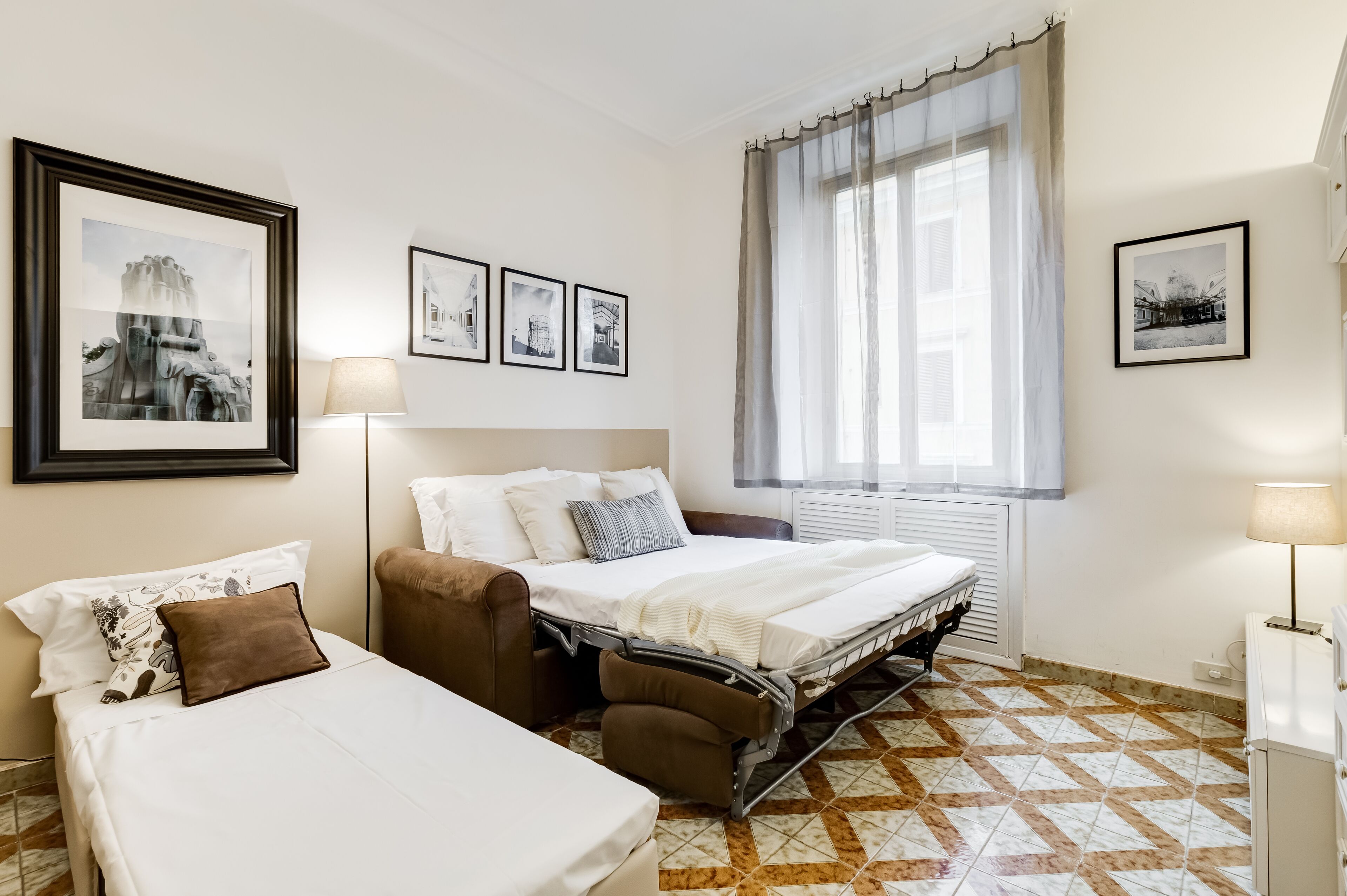 Habitación de hotel con cama, sofá, sillón y ventana con cortinas. En la pared hay cuadros enmarcados.