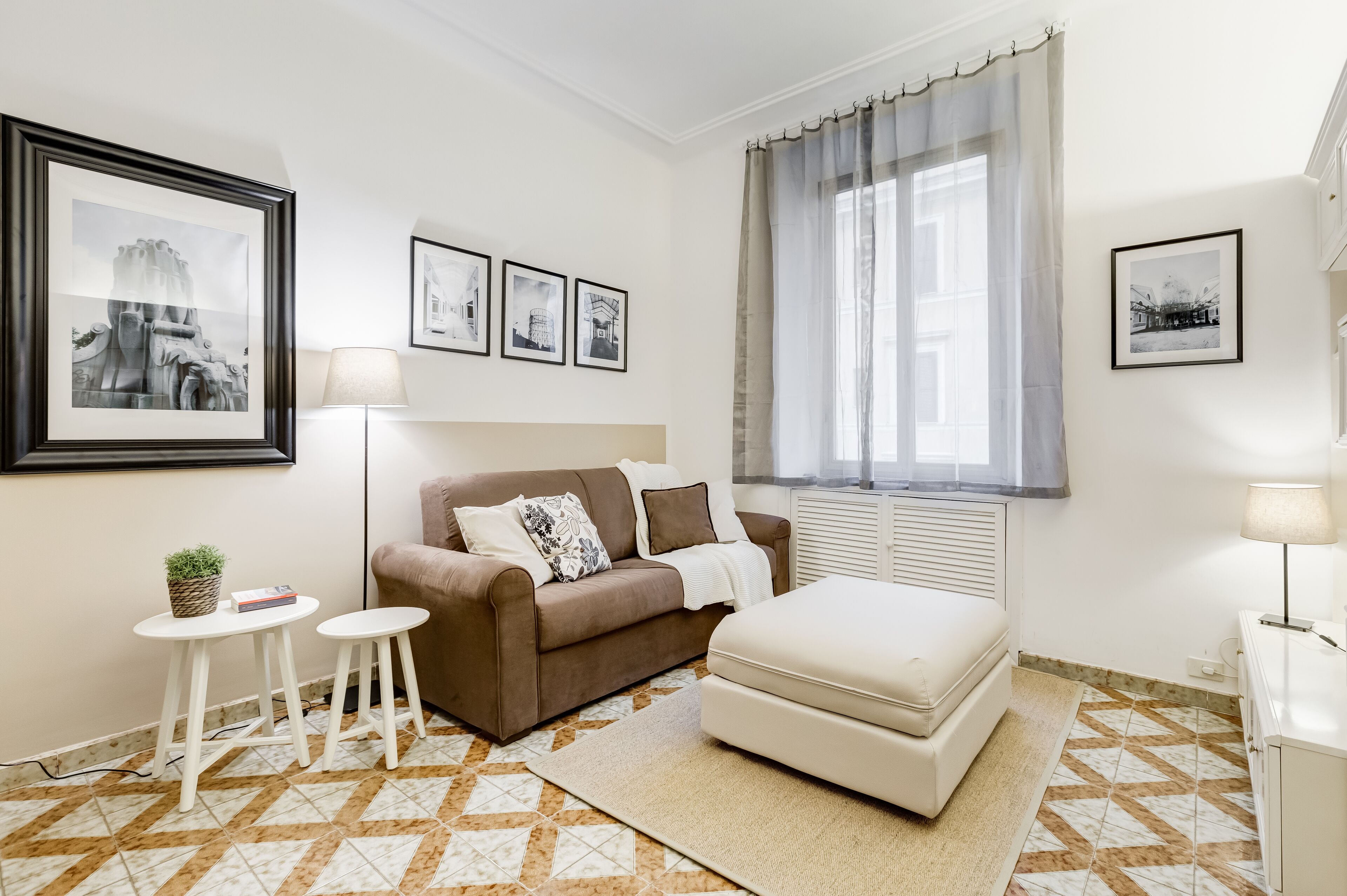 A modern living room with a brown sofa, a white ottoman, a small white table with a plant, and a floor lamp. There are framed pictures on the walls and a window with curtains.