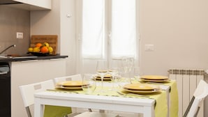 Une table à manger dressée avec des assiettes, des verres et des couverts, une horloge au mur et un panier de fruits sur le comptoir.