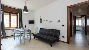 Un salon moderne avec un canapé noir, une table à manger et des chaises. Un téléviseur est fixé au mur, et un mur porte une citation inspirante.