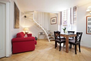 A living room with a red sofa, a dining table with chairs, a staircase, and a window with curtains.