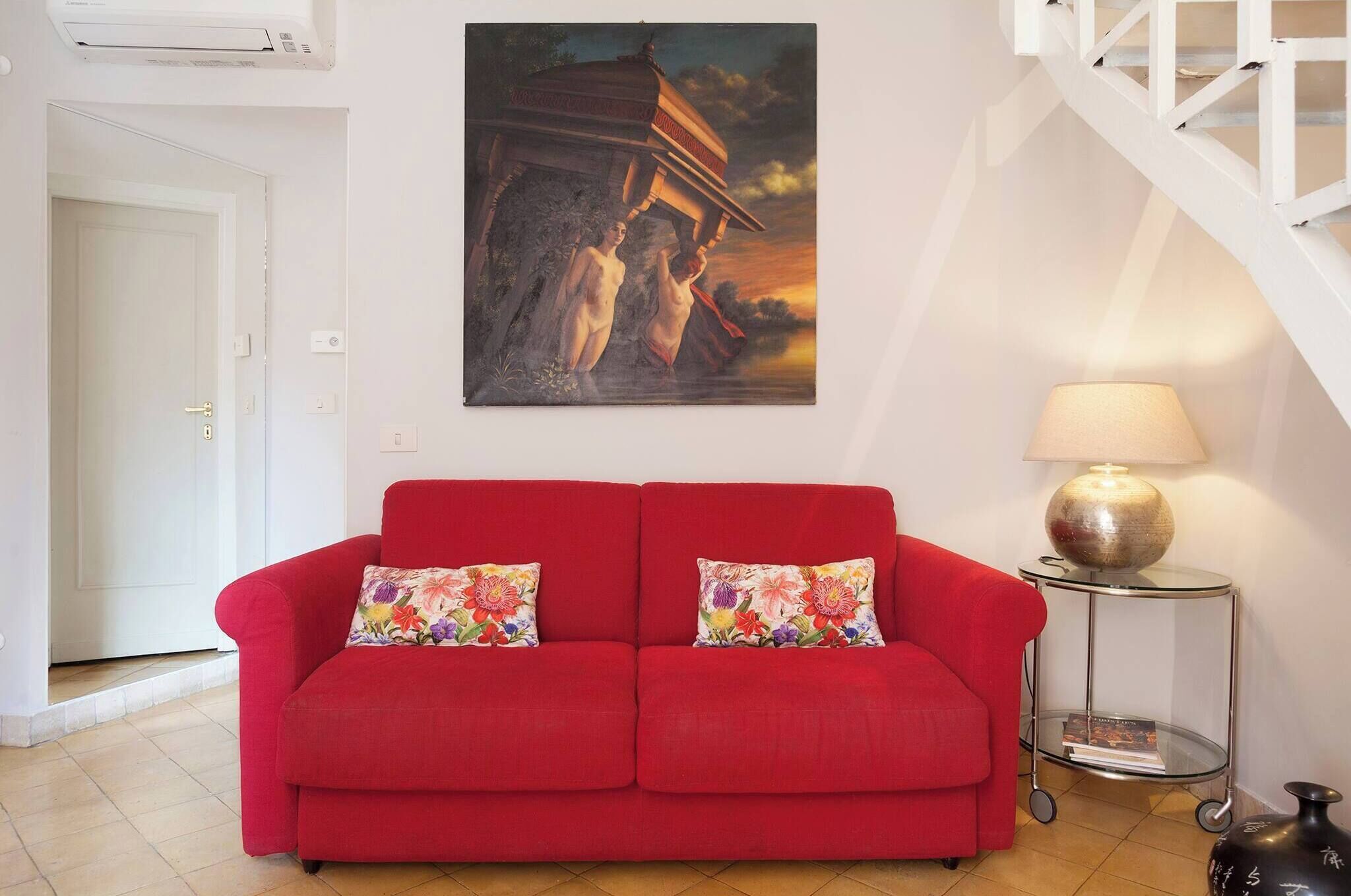 A red sofa with floral cushions, a painting of two figures on a balcony, a lamp, and a small table with a book.