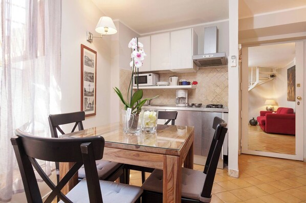 A modern kitchen with white cabinets, a microwave, and a coffee machine. There is a dining table with chairs and a vase with flowers.