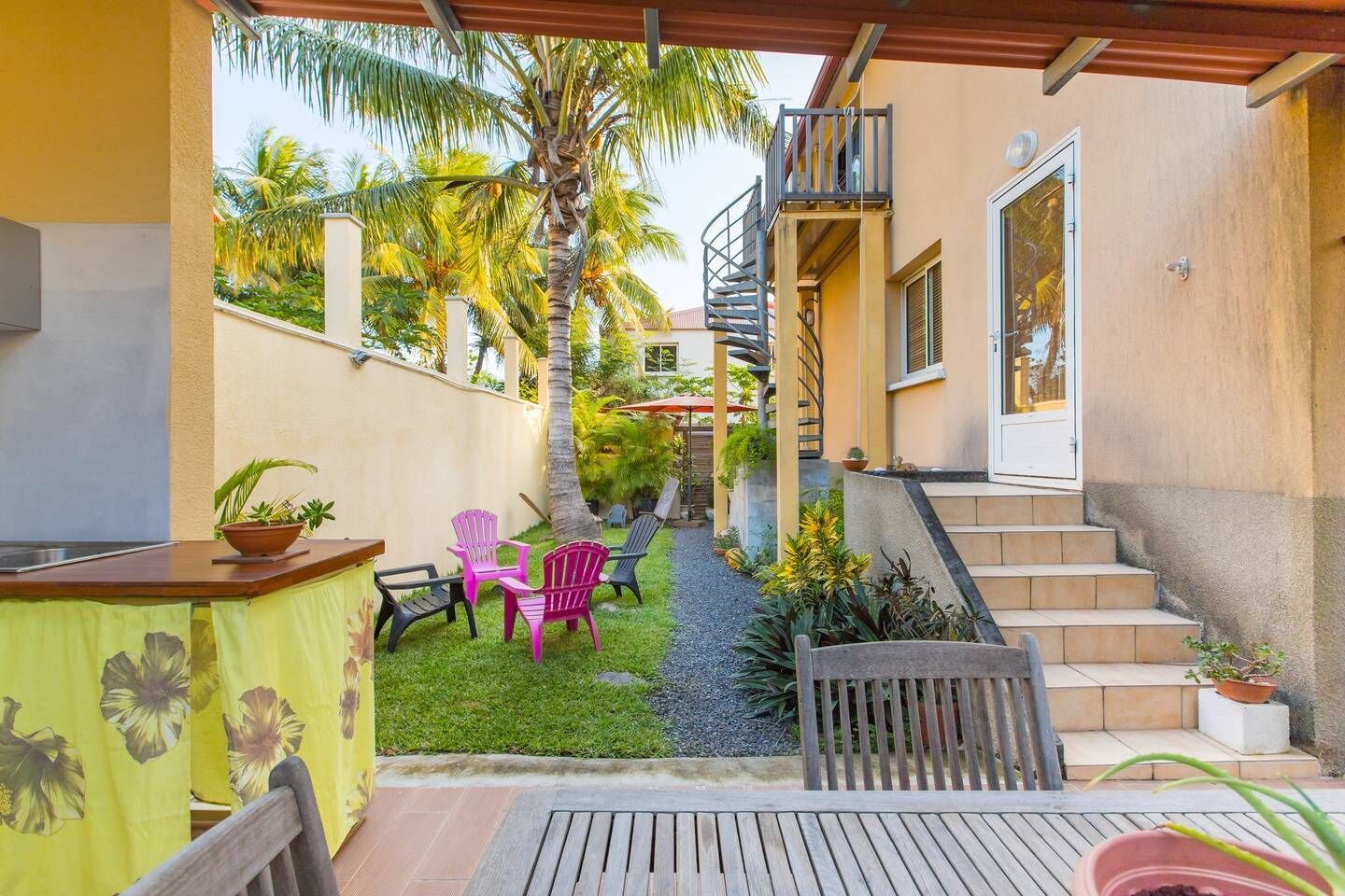 A covered outdoor area with a wooden table, chairs, and a small garden with palm trees and plants.