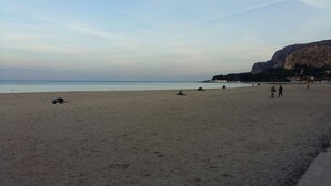 A wide sandy beach with a calm sea, a few people scattered along the shore, and a rocky cliff in the background.