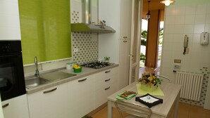 A kitchen with a sink, stove, and cabinets. There is a chair and a table with a basket of fruit.