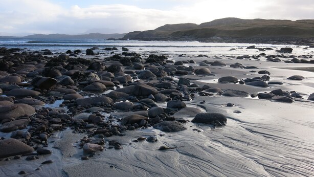 A rocky beach with waves, a clear sky, and distant hills.