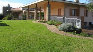 Ein zweistöckiges Haus mit überdachter Terrasse, Steinsäulen und einem gepflegten Rasen.