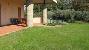 Eine überdachte Terrasse mit Steinpfeilern, einer Sitzbank und einem Garten mit verschiedenen Pflanzen und einem Zaun.