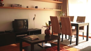 Een woonkamer met een televisie, een salontafel met tijdschriften en een eethoek met rieten stoelen en een tafel.