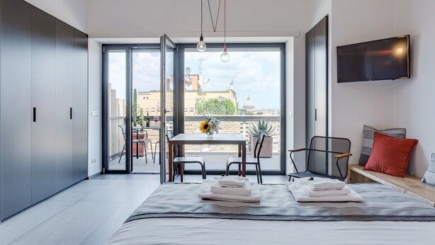 A modern bedroom with a large bed, a TV mounted on the wall, and a balcony with a table and chairs. - Studio Vaticano Int. 5B - Terrace with View on San Pietro (Roma)